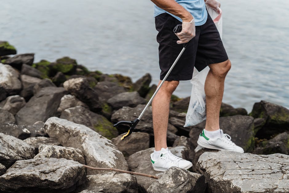 A person dressed in casual outdoor clothing, wearing a light blue T-shirt, black shorts, and white sneakers with green accents, stands on a rocky shoreline near a body of water. The individual is hold