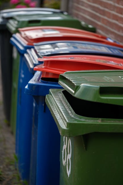 A person’s pale arm extends into the frame, holding a green plastic rubbish bag filled with waste. The bag appears to be full and tightly sealed at the top with a knotted twist tie, with some transp