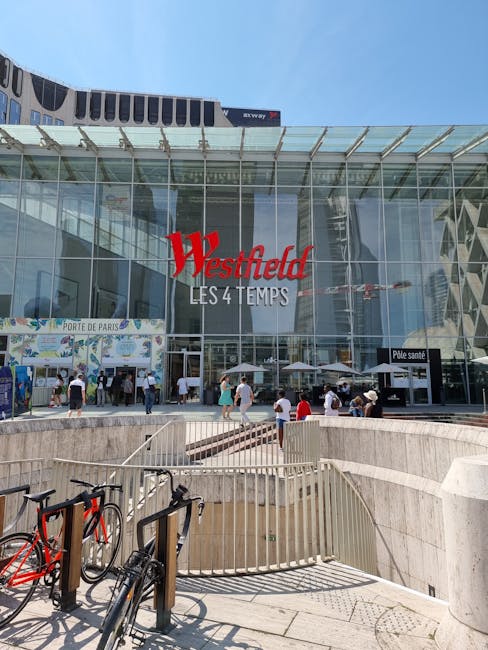The image depicts the entrance to Westfield London shopping centre, with a large glass facade reflecting the surrounding urban environment. Prominent red signage displays the name 'Westfield LES 4 TEM